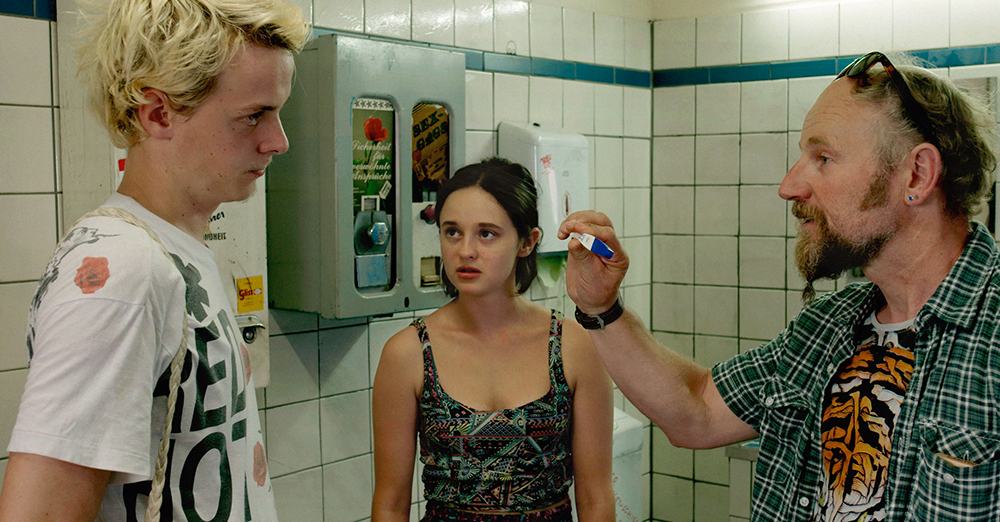 Three people in a motorway toilet. Condom and tampon dispenser are behind them on a white tiled wall. On the right side, a middle aged man is showing a pregnancy test. The young woman in the middle is looking expectantly at the young man on the left.