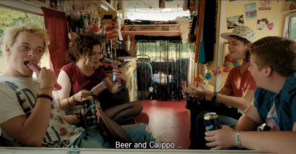 The four young band members are sitting in a caravan called Bertl. They are drinking beer and eating Calippo popsicles. The caravan is decorated with fake flower garlands, post cards and a tinsel curtain. On the kitchen counter, there is a pizza box and empty food packaging.