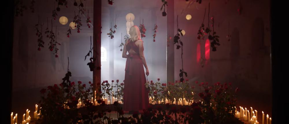 Wide shot of singer Nele in a red dress in a hall. She is standing with her back to the camera, in a circle of burning candles and red roses. The hall is dimly lit, with black and red being the dominant colors.
