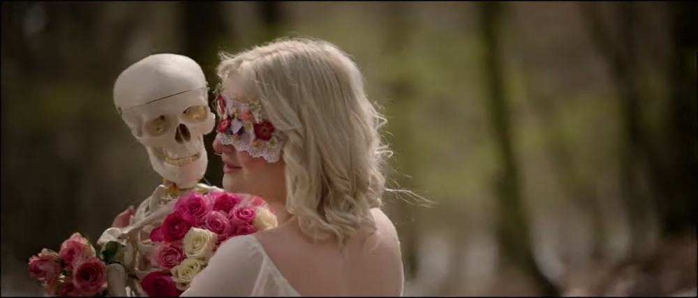 Closeup shot of singer Nele in a white dress dancing with a plastic skeleton in a forest. It is sunny. The skeleton is covered in white and pink roses. A mask made of pink lace and pressed flowers is covering Nele’s face.