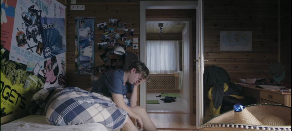 Protagonist Michael is sitting on the edge of the bed of his teenage bedroom. The walls are covered in wooden panels and decorated with photos and posters. The door is open, the opposing bathroom is partially visible. Sleepy-eyed Michael is covering his face with his hand.