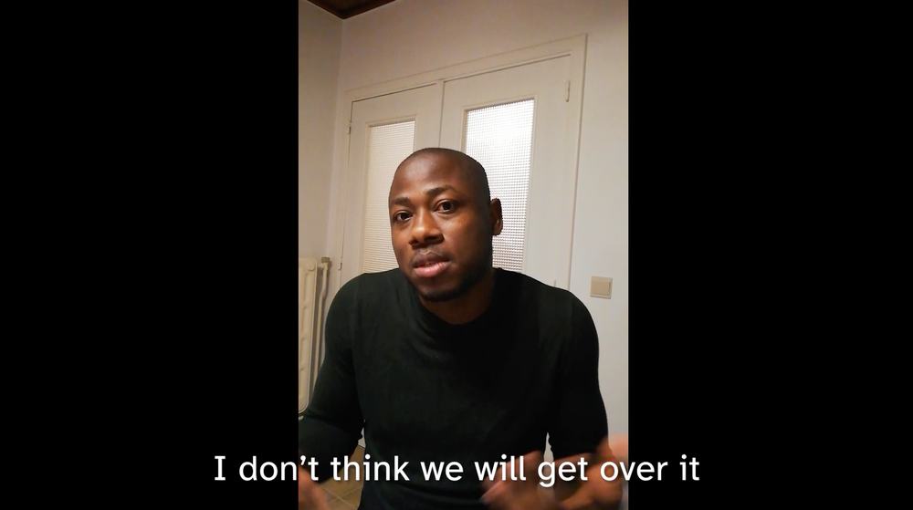 Protagonist Aaron is sitting in a hotel room. He is wearing a black sweater. His shoulders are pulled up and he is looking directly into the camera.