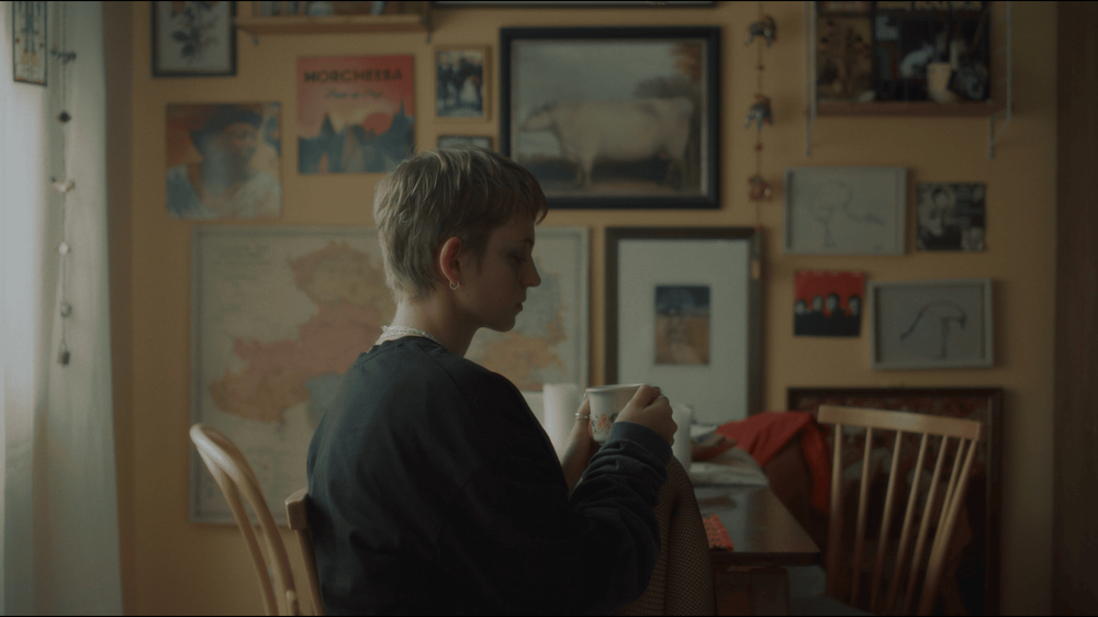 The protagonist sitting at a table, showing her side to the camera. She is holding a cup in her hand. The wall behind her is painted peach and decorated with a mix of colorful artworks.