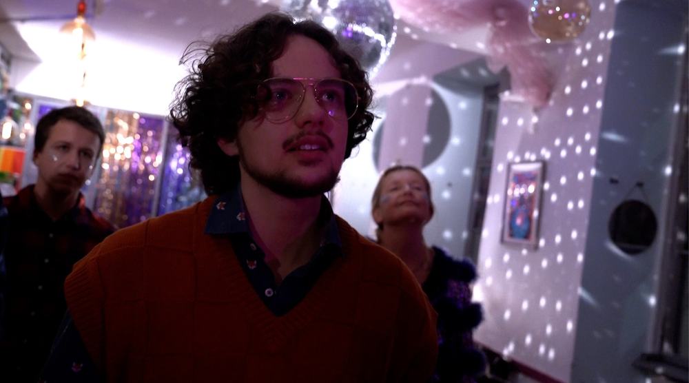 Closeup of main character Noah. He has wavy brown hair and glasses. He is standing in a queer café, behind him two people. A disco ball scatters light onto the white walls. Noah is looking straight ahead in an emotional way, but is also lost in thought.
