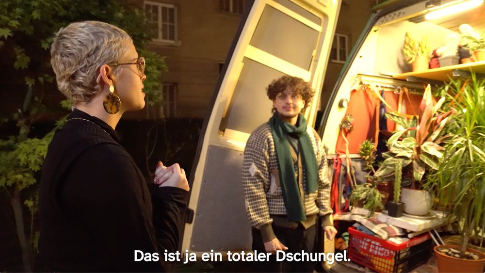 Supporting character Miriam is standing in a yard together with Noah. The back doors of Noah’s van are open. The van is filled with plants and boxes. The subtitles translate to: “That’s a total jungle.”