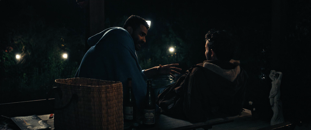 The two main characters in conversation on a terrace. It is night. Next to them wine bottles and a bag. Beyond them, a pond and a garden that is dappled with light bulbs.