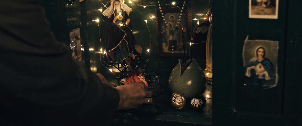 Closeup shot from behind of the grandmother placing flowers in a little shrine. The shrine is open and decorated with Christian images and a statue of the Virgin Mary. Incense sticks are burning. It is illuminated by fairy lights.