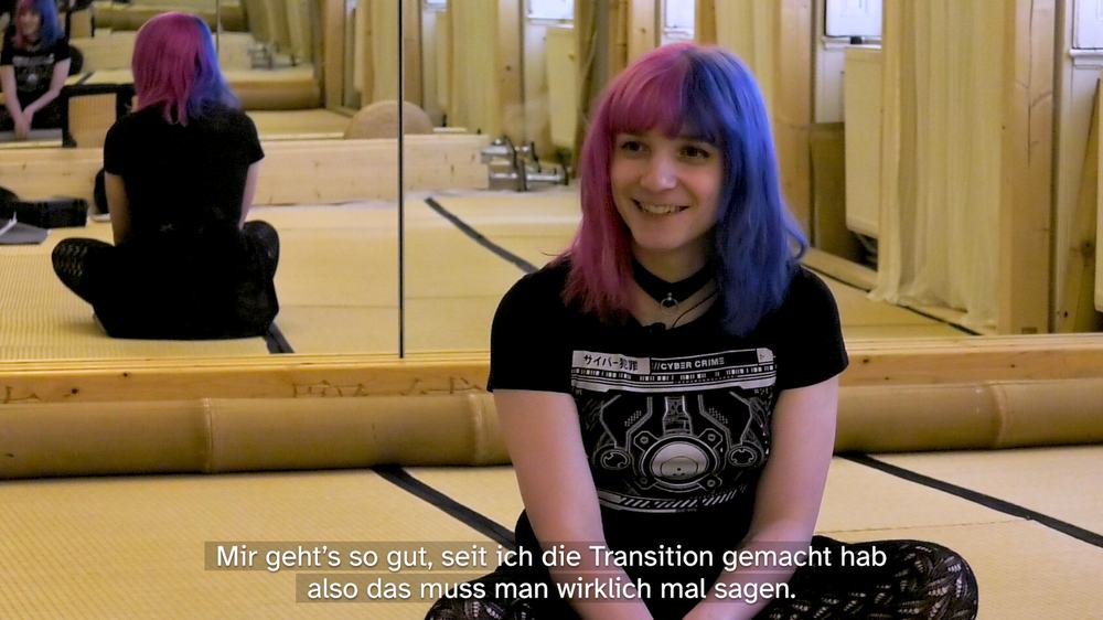 Protagonist Mia is sitting cross-legged on mats of a rope-play studio. She has blue and pink hair and is smiling. The mirrored wall behind her shows her mirrored images a few times. The subtitles translate to: “I am feeling so good since I transitioned, I really need to say it.”